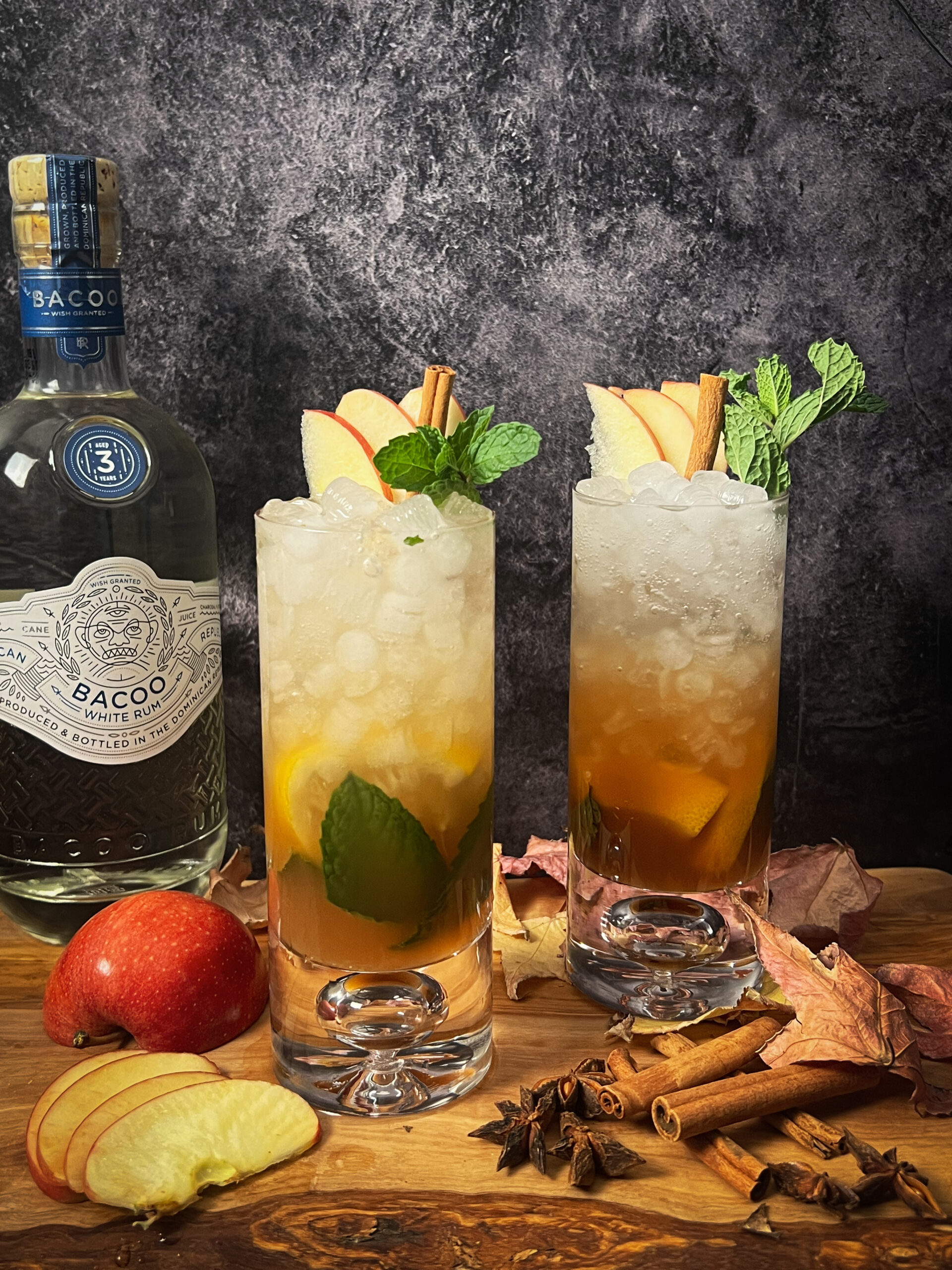 2 apple cider mojito cocktails on a wood block with a bottle of BACOO Rum